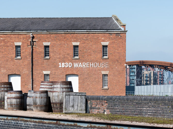 End view of the 1830 Warehouse showing its bold painted name, with barrels and cobbled surroundings reflecting its historic role linking rail, road and canal trade.
