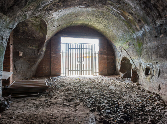 Interior view of a brick-lined tunnel chamber ending at a metal gate, with rough ground and exposed brickwork showing wear and age.