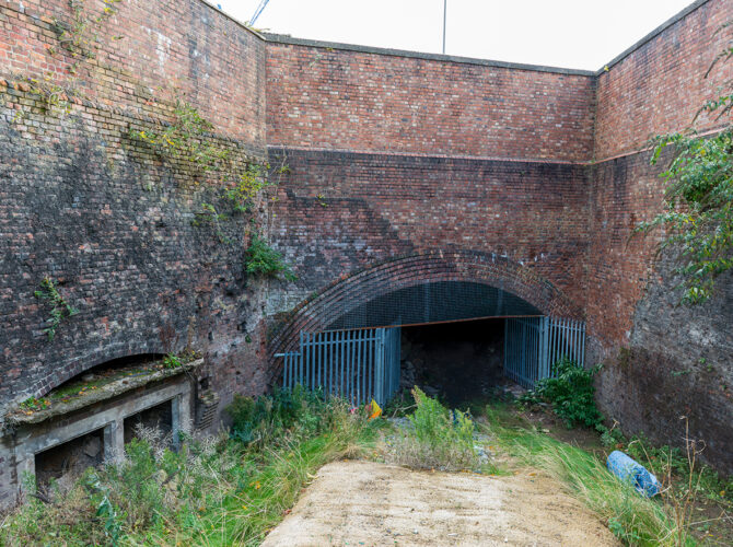 View into a brick-lined cutting with a single arched tunnel entrance secured by metal fencing, surrounded by vegetation and weathered walls.