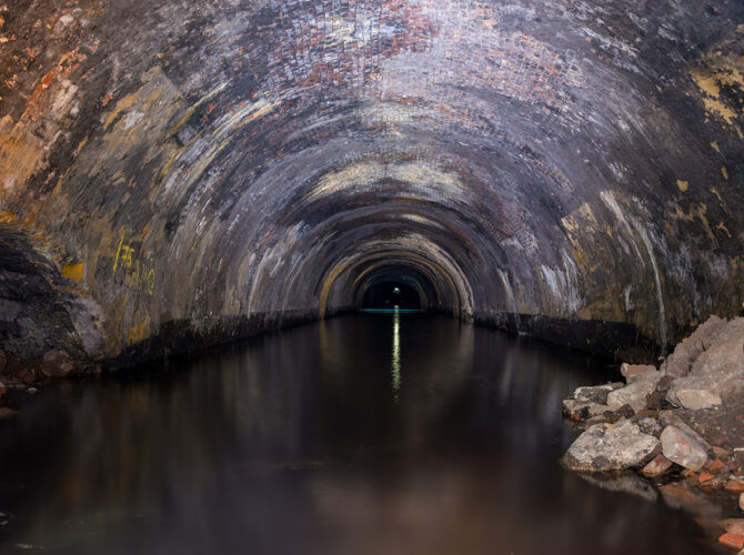 Central view inside the Wapping Tunnel, showing the curved brick lining reflected in still water, with rubble at the edge and a distant point of light ahead.