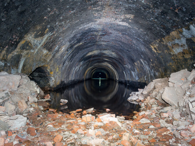 View into the Wapping Tunnel from a debris-strewn entrance, leading into a partially flooded brick-lined passage.