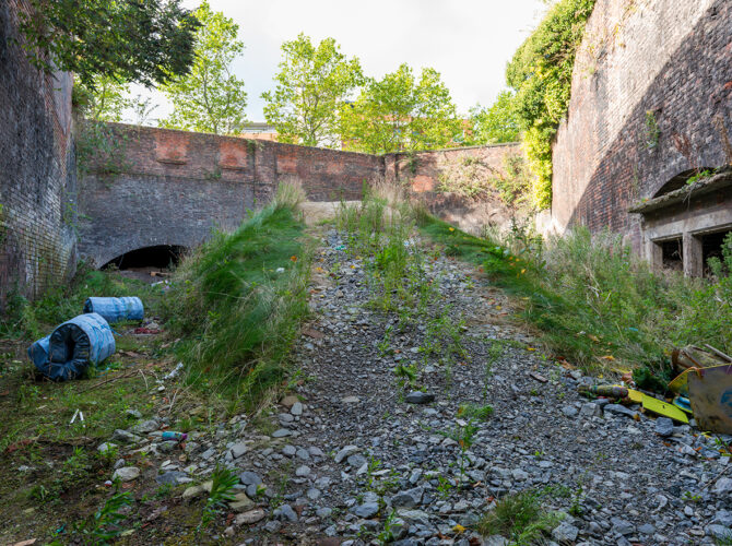 Wide view of a disused cutting filled with rubble and grass, leading toward dark tunnel openings beneath high brick retaining walls.
