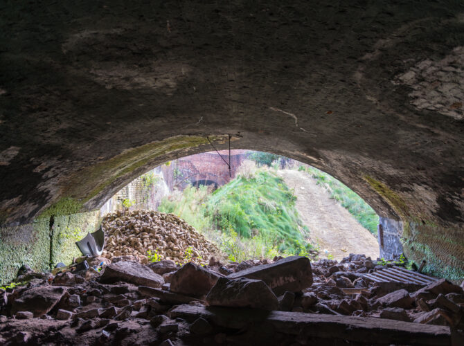 Perspective from inside a brick arch tunnel looking outward to daylight, with piles of stone and debris in the foreground and vegetation beyond.