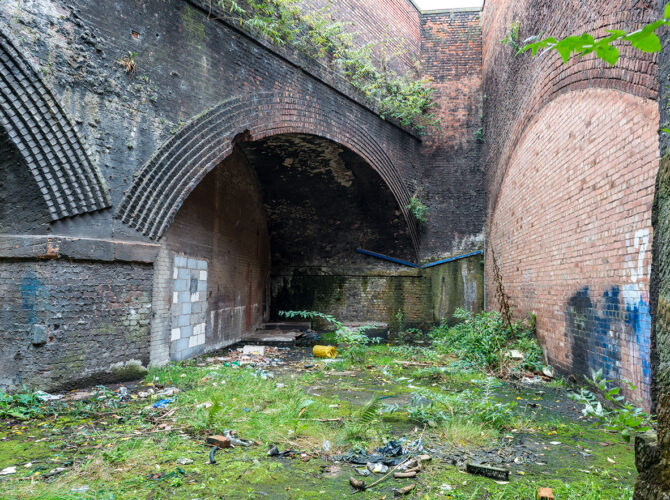 Exterior view of a large arched opening leading into the goods station, surrounded by moss, plants, and discarded materials.