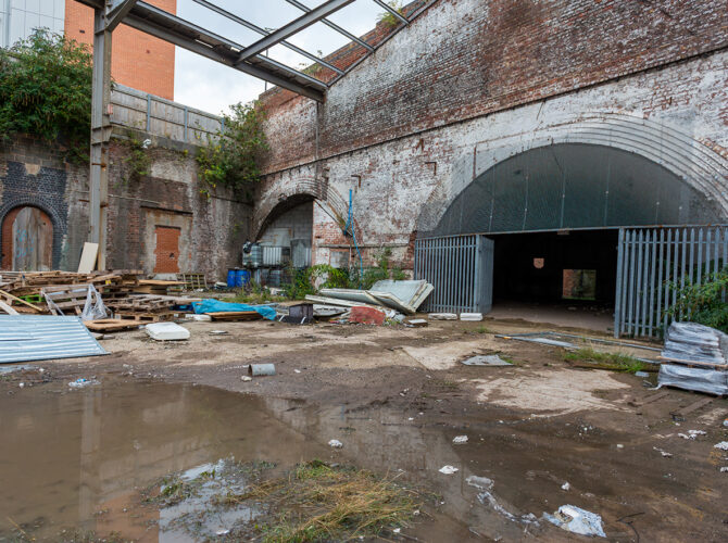 View of a cluttered yard with puddles and construction debris in front of large brick arches and a gated tunnel entrance.