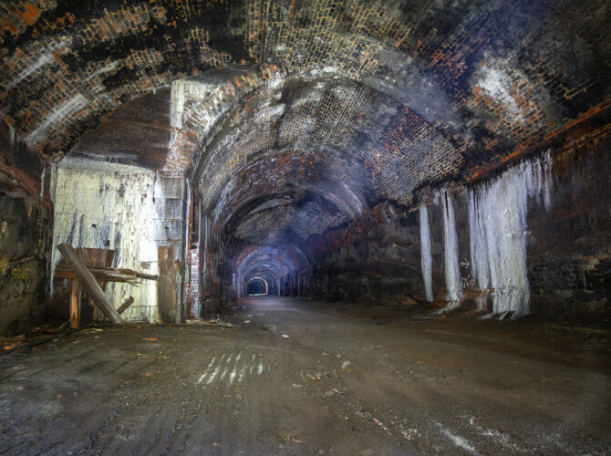 Deep view along the Wapping Tunnel, with mineral staining on the brick walls and a distant point of light marking the far end.