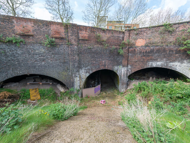 Three adjacent arched openings set into a high brick wall, with vegetation and debris in the foreground and one entrance partially blocked.