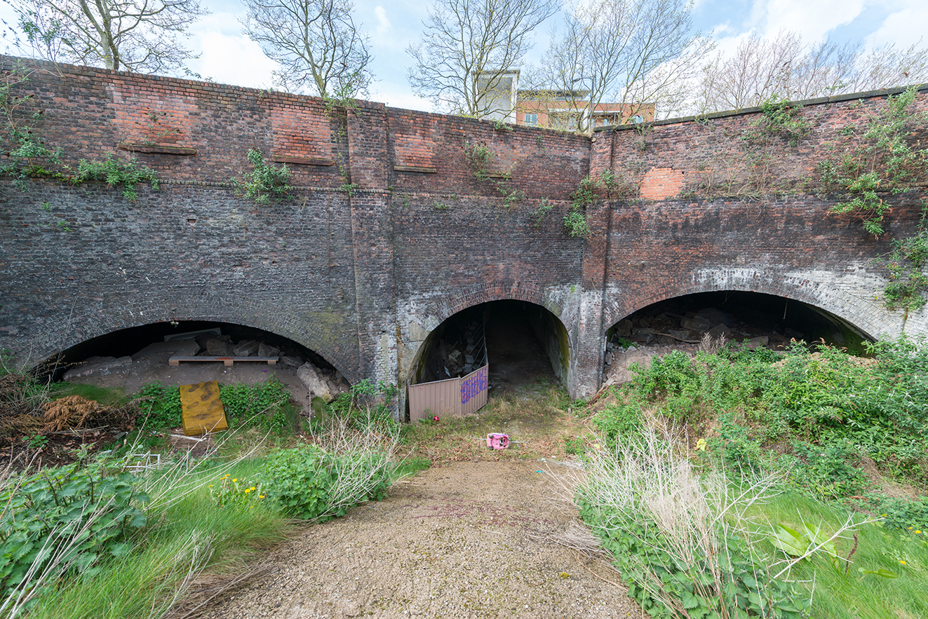 Three adjacent arched openings set into a high brick wall, with vegetation and debris in the foreground and one entrance partially blocked.