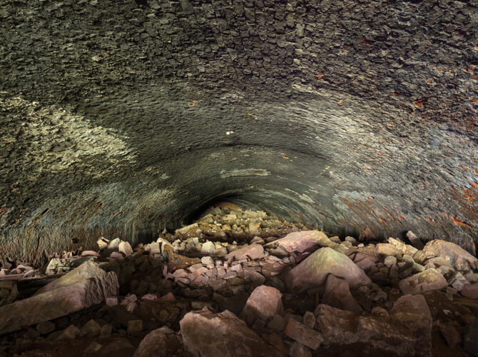 Interior view of a narrow tunnel filled with rocks and debris, showing the curved brick lining and dim, enclosed space.