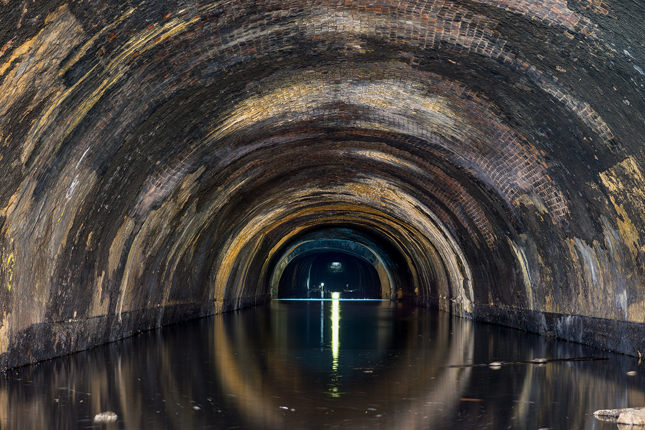 Inside the Wapping Tunnel near Crown Street, showing the brick-lined arch reflected in still water with a faint light in the distance.