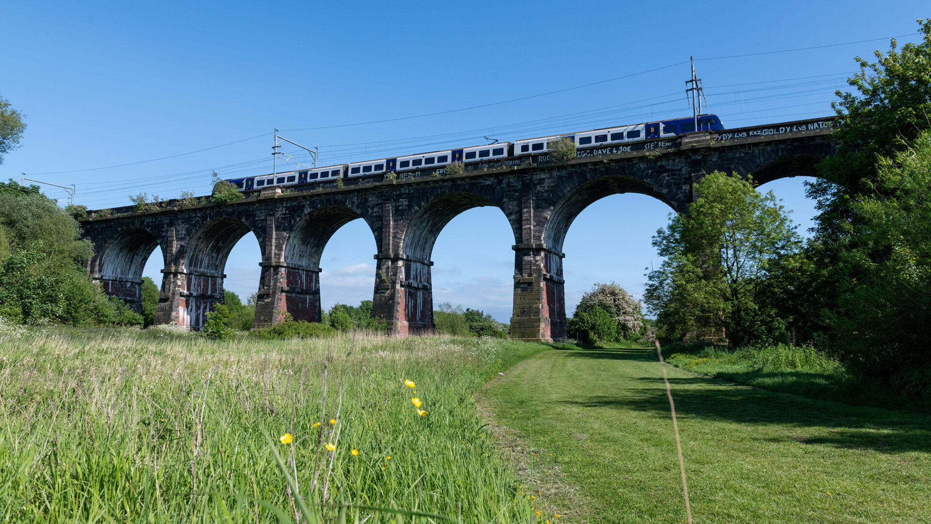 A modern passenger train travels across the historic arches of Sankey Viaduct high above the valley.