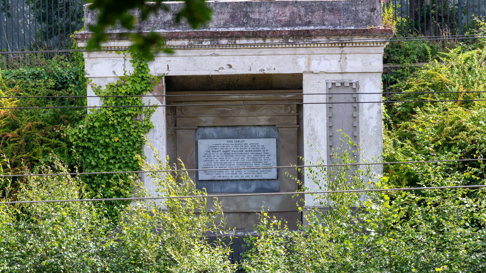 The classical-style Huskisson Memorial set into the railway cutting, partly hidden by trees and vegetation, with the commemorative plaque visible at its centre.