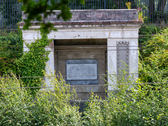 The classical-style Huskisson Memorial set into the railway cutting, partly hidden by trees and vegetation, with the commemorative plaque visible at its centre.