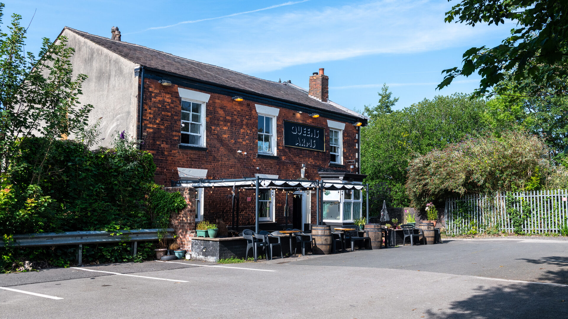 A view of the Queens Arms from the car park showing the two-storey red-brick building, outdoor tables and surrounding trees.