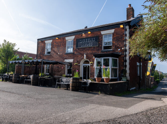 The red-brick Queens Arms building with outdoor seating, canopy and the pub sign above the entrance