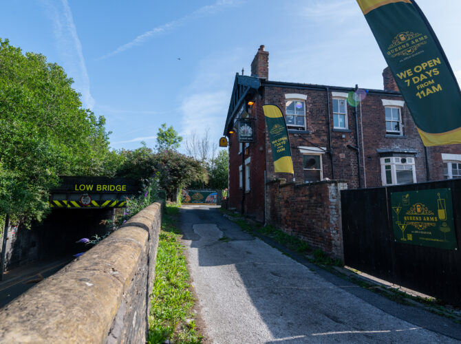 The Queens Arms pub seen from the nearby lane, with railway bridge signage and the building’s exterior flags and signs visible