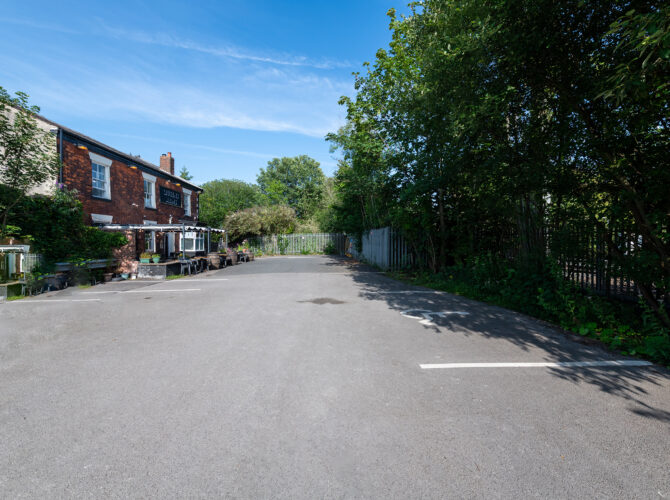 A wide view of the car park outside the Queens Arms pub, with the red-brick building on the left and trees and railway fencing along the edge of the site.