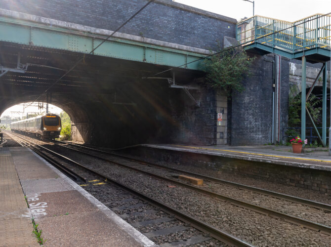 Passenger train passes under the bridge at Eccles station. The station platform, overhead electrification and metal footbridge stairs are visible beside the tracks.