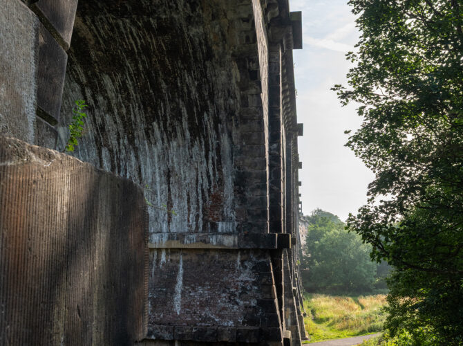 A path passes beneath the towering stone piers of the viaduct, framed by trees and greenery.