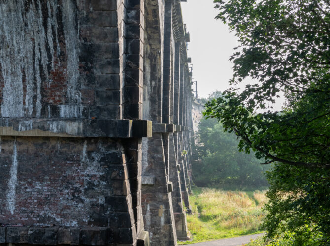Looking along the underside of the viaduct where a path runs beside the stone piers and trees.