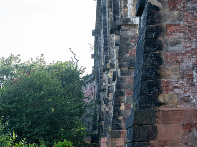 A side view showing the repeating sandstone piers and brickwork of the viaduct rising above vegetation