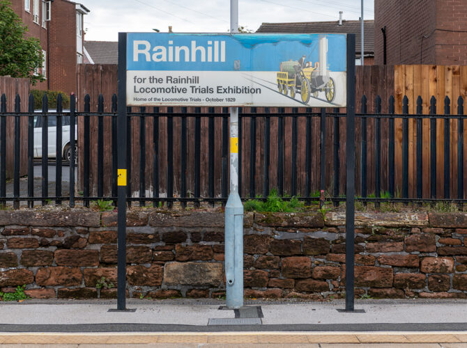 Station sign highlighting Rainhill as the site of the 1829 Locomotive Trials, mounted in front of fencing and houses.