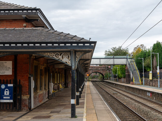 View along a platform with a decorative canopy, looking towards a footbridge and railway bridge in the distance.