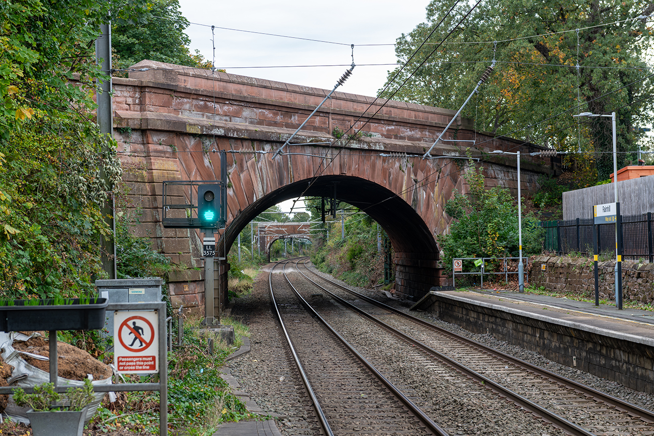 Close view of the sandstone Skew Bridge spanning the railway, with tracks curving beneath and overhead lines above.
