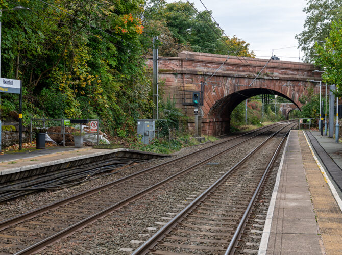 View along the tracks at Rainhill station looking towards a large sandstone bridge, with trees and platform signage on either side.