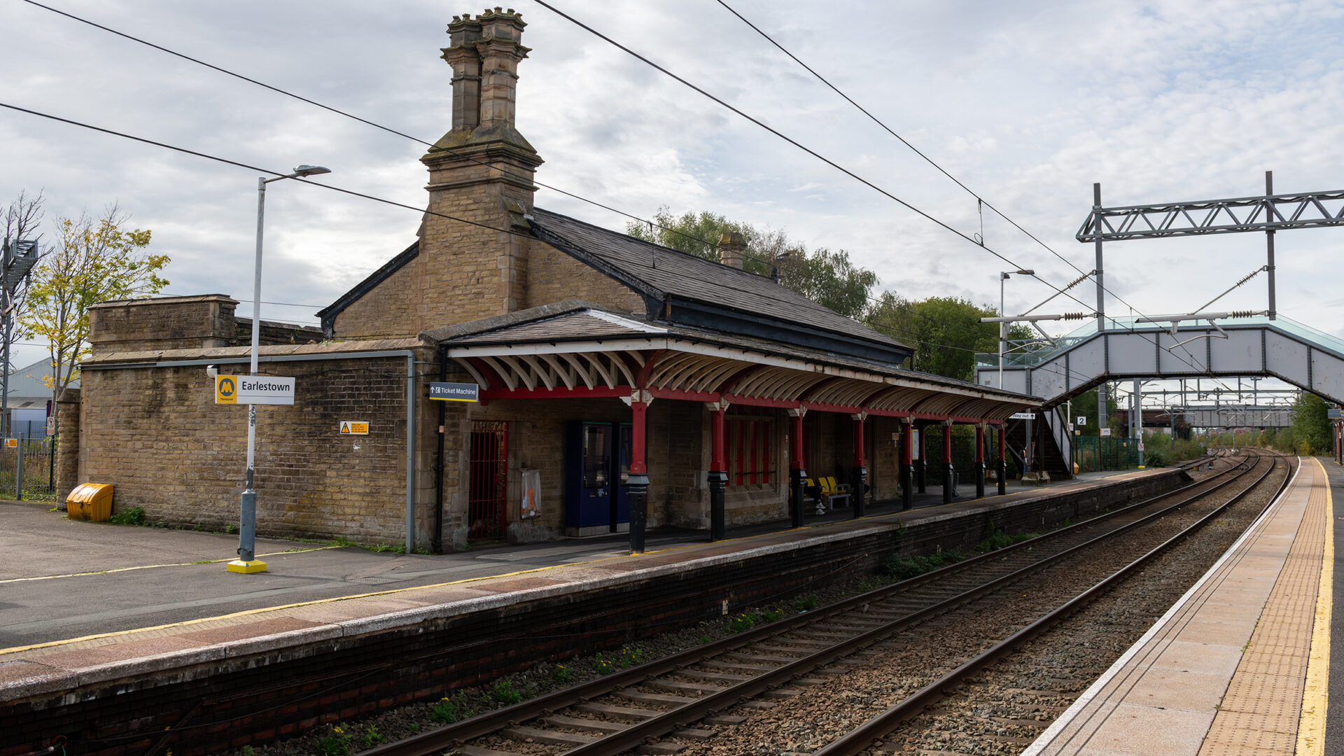 A wider view of Earlestown Station showing the historic station building, platform canopy, railway tracks and footbridge in the distance.