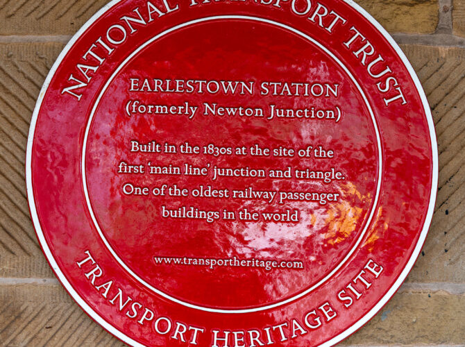 A red National Transport Trust plaque marking Earlestown Station as a transport heritage site and noting its connection to the first main line railway junction.