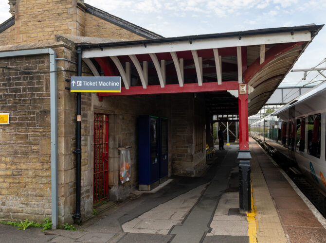 A Victorian station building and red-painted timber canopy seen along the platform, with a train standing on the track