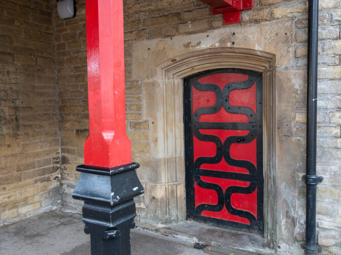 A close view of a red and black decorative door set into the stone wall of Earlestown Station beneath the painted timber platform canopy.