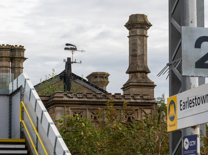 A Merseyrail Earlestown station sign stands in the foreground while the historic station building with decorative chimneys and weather vane rises behind it