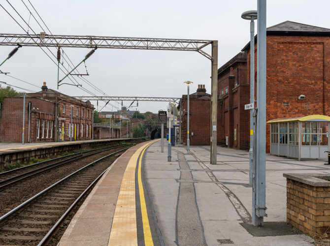 Platform view looking down the line towards the tunnel, with overhead wires, historic buildings and tracks curving into the distance.
