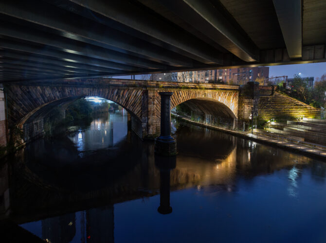 The bridge illuminated at night beneath a road bridge, with warm lighting highlighting the arches and reflections in the river.