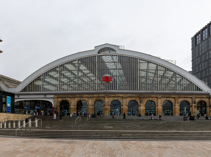 Frontal view of the station’s curved glass train shed and arched entrance, with passengers moving across the forecourt steps.