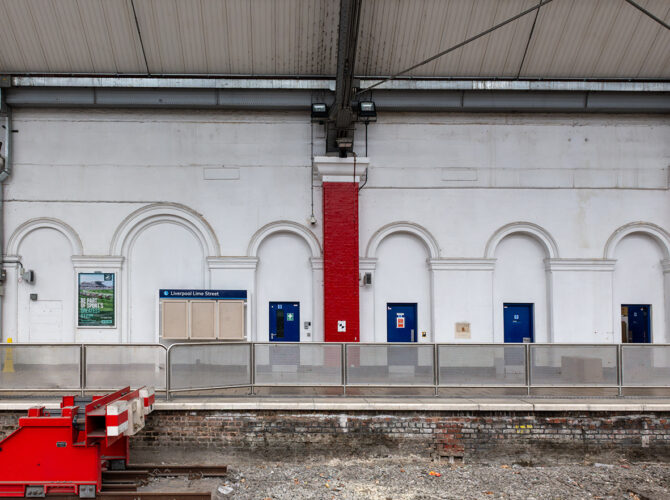 Interior platform view showing a white wall with decorative arches, blue doors, and red columns behind a safety barrier.