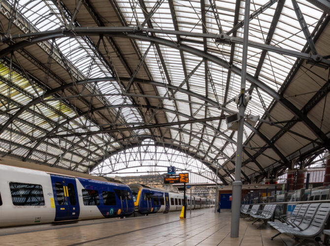 View along a platform with trains waiting beneath the large iron and glass roof structure, with seating and signage visible.