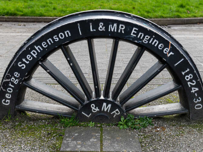 Close-up of a black metal wheel engraved with “George Stephenson L & MR Engineer 1824–30,” displayed as a heritage feature outside the station.