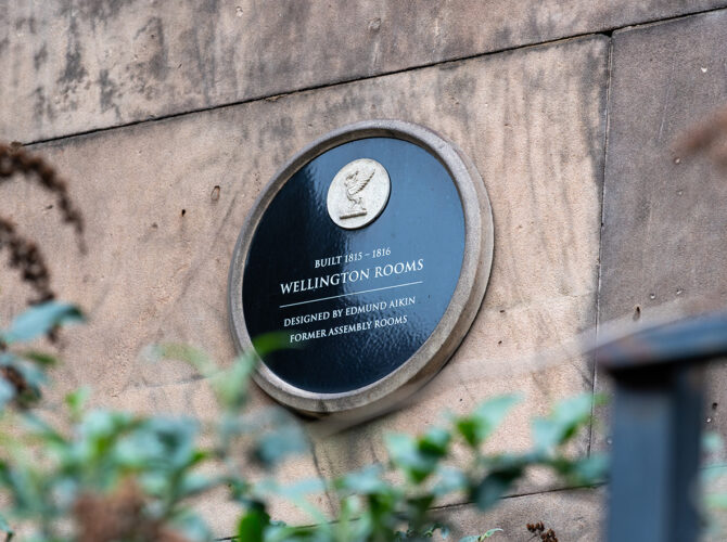 Close-up of a circular plaque on the stone wall noting the Wellington Rooms, built 1815–1816 and designed by Edmund Aikin.