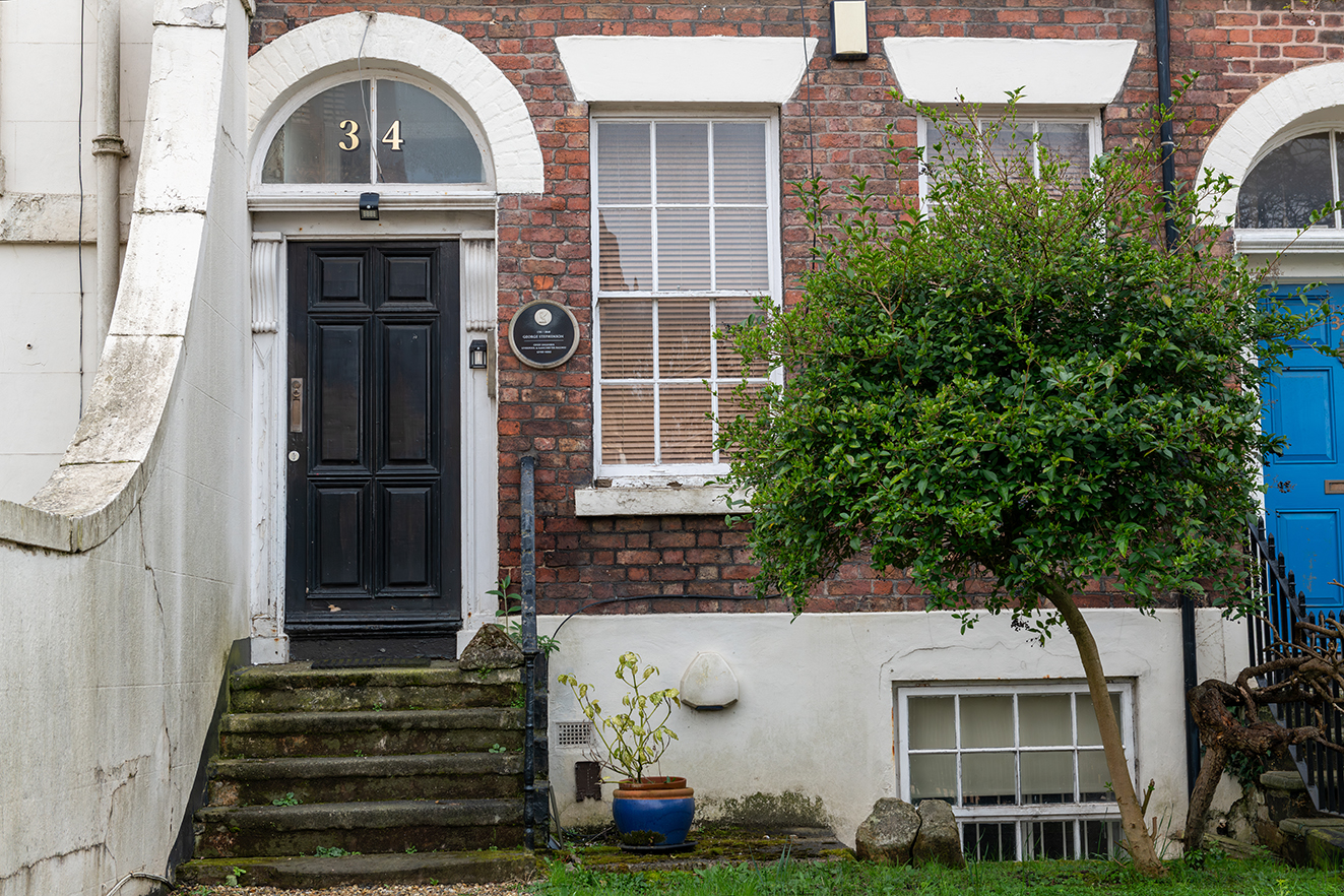 A brick townhouse with steps to a black front door, featuring a plaque marking George Stephenson’s residence.