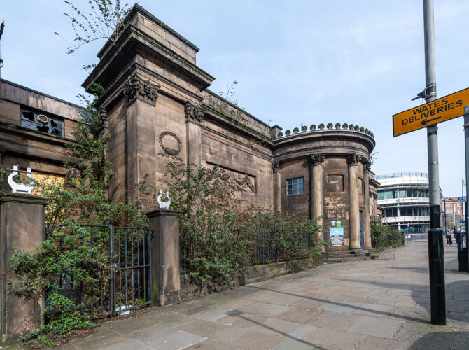 The weathered stone building sits behind railings and shrubs, with classical detailing and a quiet city street beside it.