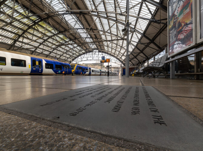 Trains stand under the iron roof, with a plaque in the foreground marking the station’s railway heritage.