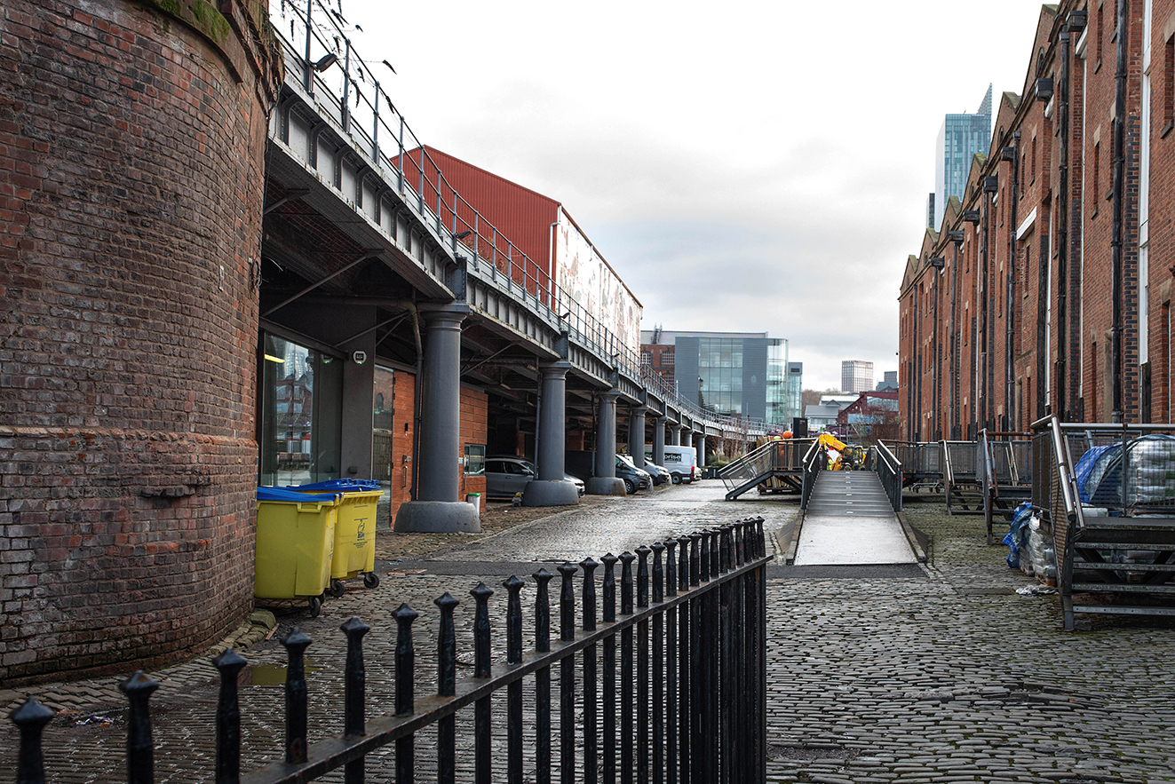 View along a cobbled service road beside the goods depot, showing a brick viaduct carried on a series of arches and columns, with railway structures extending overhead.
