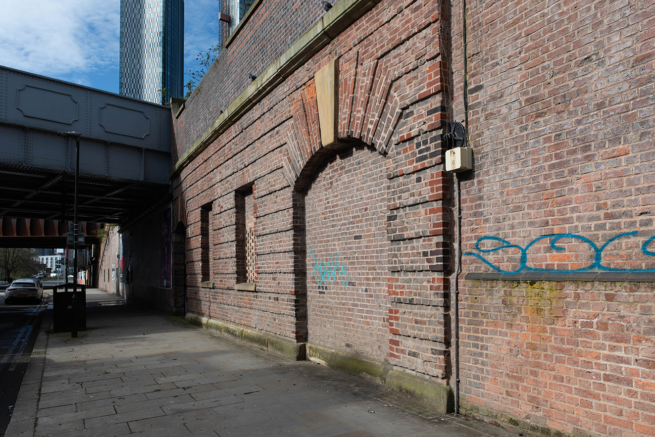 Original red-brick arrivals station wall with infilled arches and later alterations under a modern bridge.