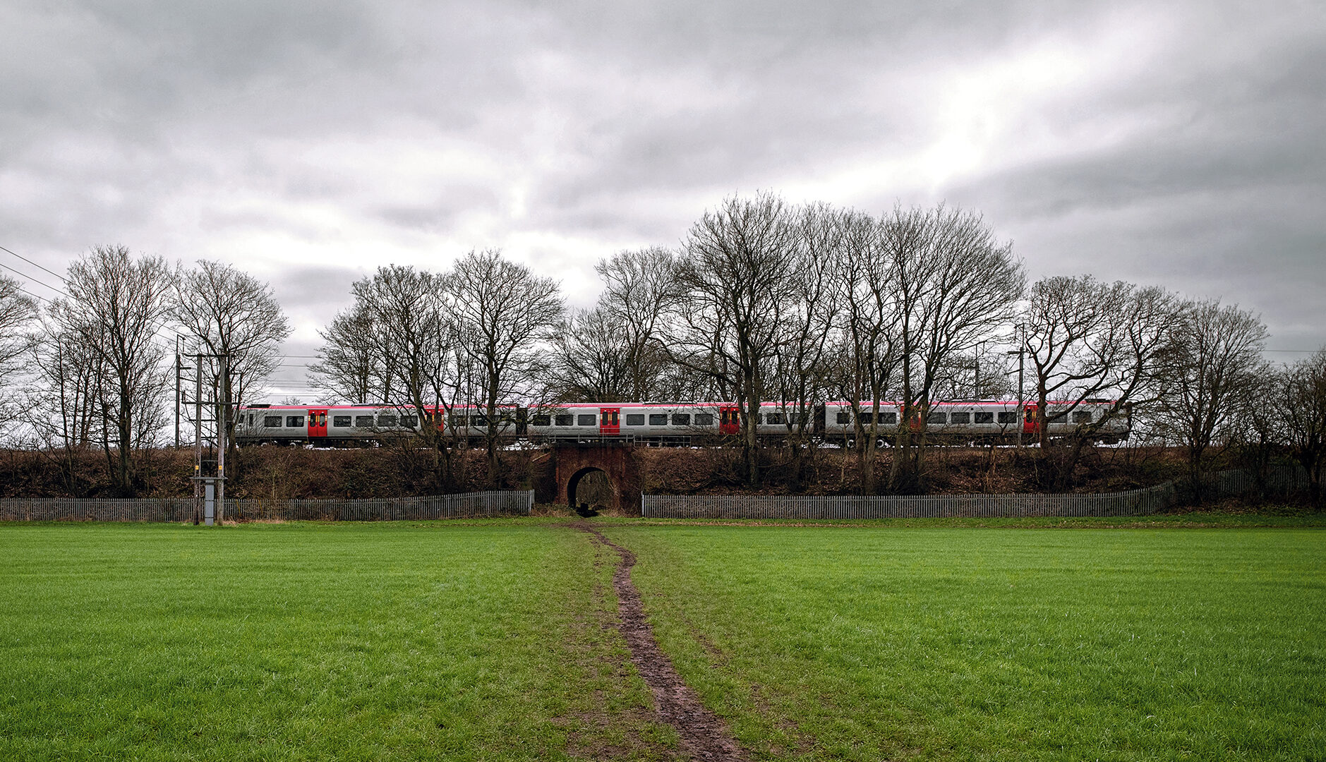 Passenger train crossing a small brick railway bridge over a muddy footpath on Chat Moss.
