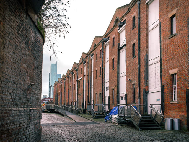 Perspective view along the former Liverpool Road warehouse range, showing repeated brick bays with loading openings, shuttered doors, and metal steps facing the cobbled yard.