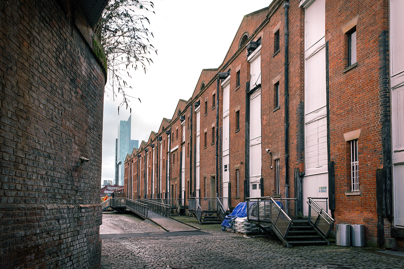 Perspective view along the former Liverpool Road warehouse range, showing repeated brick bays with loading openings, shuttered doors, and metal steps facing the cobbled yard.