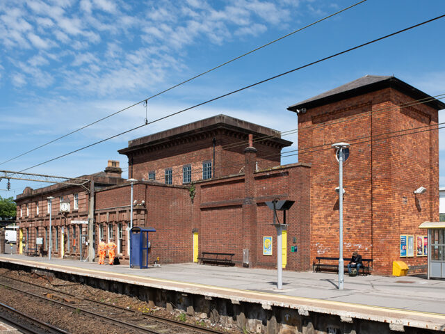 View of the brick-built hydraulic plant house and tall accumulator tower at Edge Hill Station, standing beside the platform with historic station buildings stretching behind.